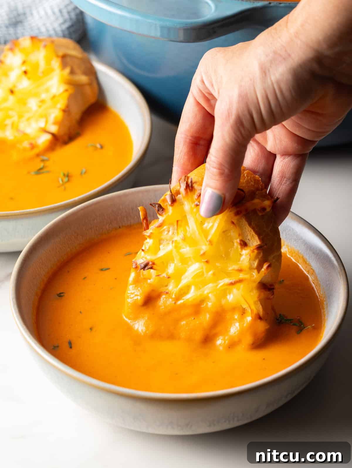 A hand holding cheesy Gouda baguette toast, freshly baked, dipping it into a bowl of vibrant, creamy roasted red pepper soup.