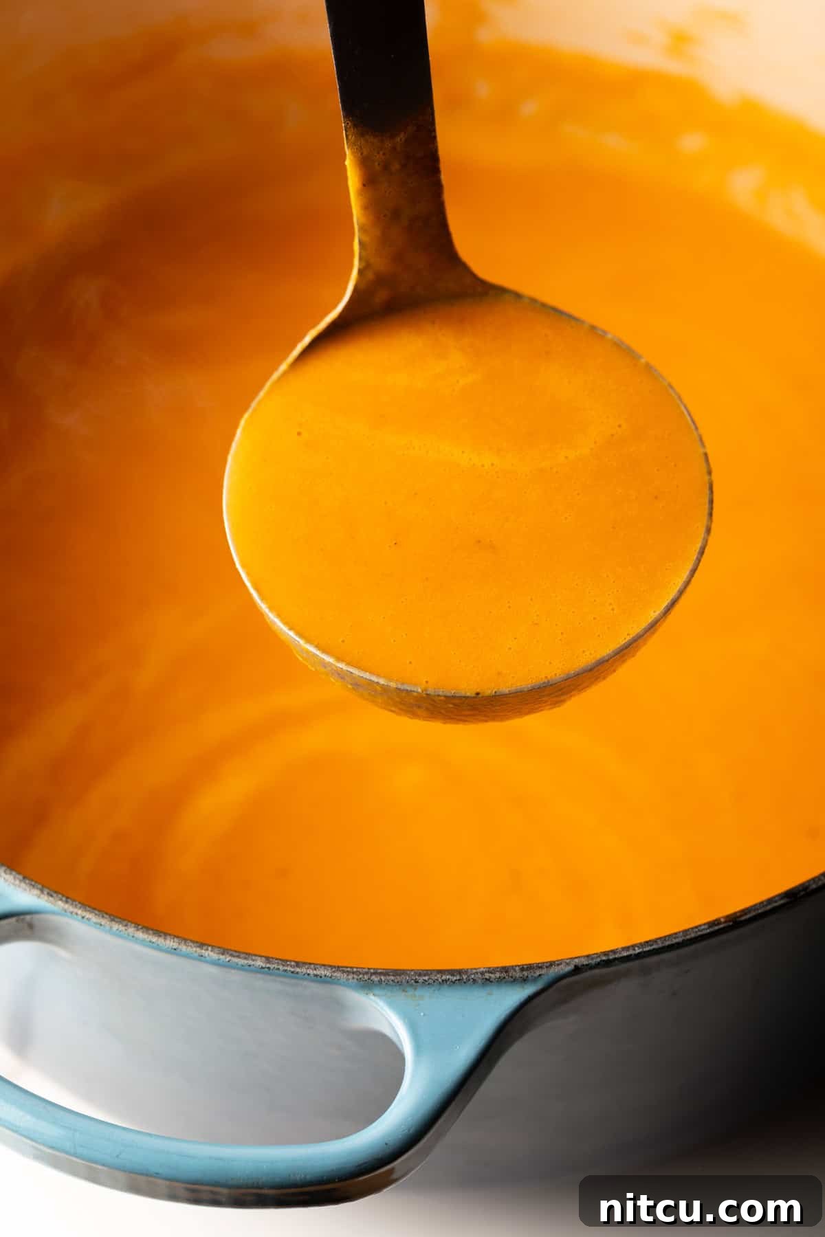 A ladle filled with rich, creamy roasted red pepper Gouda soup, held up to the camera to showcase its smooth and inviting texture.