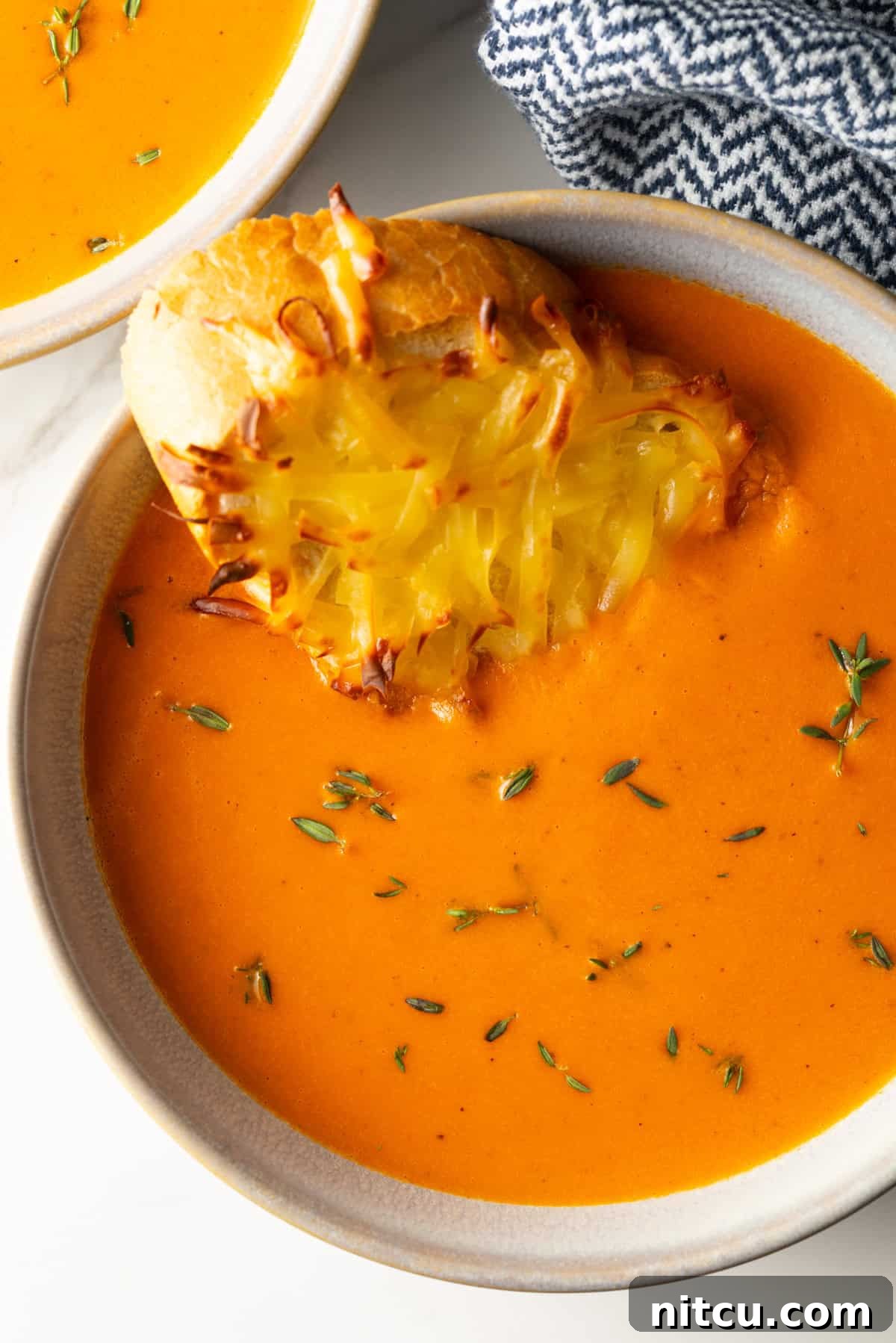 A close-up of a bowl of creamy roasted red pepper Gouda soup, garnished with fresh herbs and a slice of golden-brown cheesy baguette toast, ready to be enjoyed.
