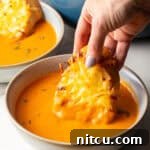 A hand holding cheesy bread that is being dipped into a bowl of roasted red pepper soup.