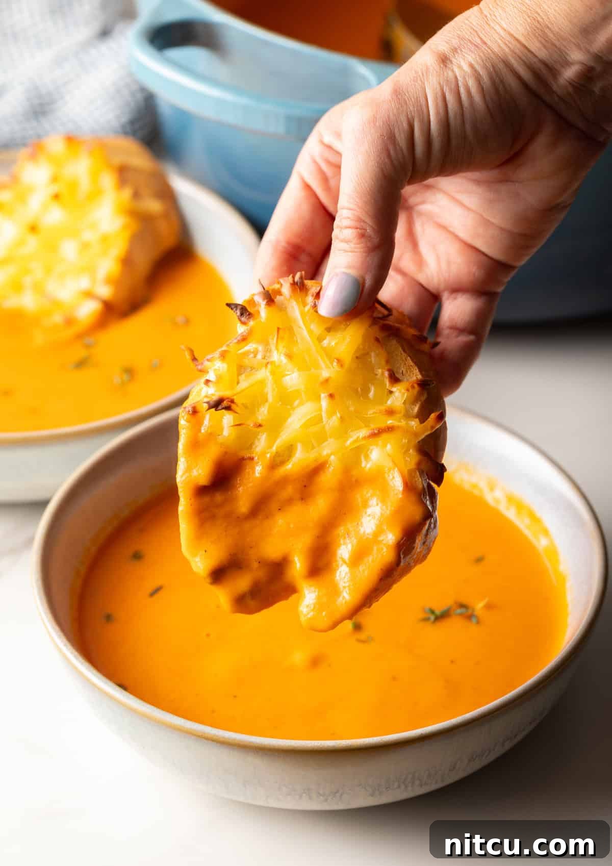 A close-up shot of a hand dipping a piece of bubbly, golden Gouda cheese toast into a rich, creamy bowl of roasted red pepper Gouda soup.