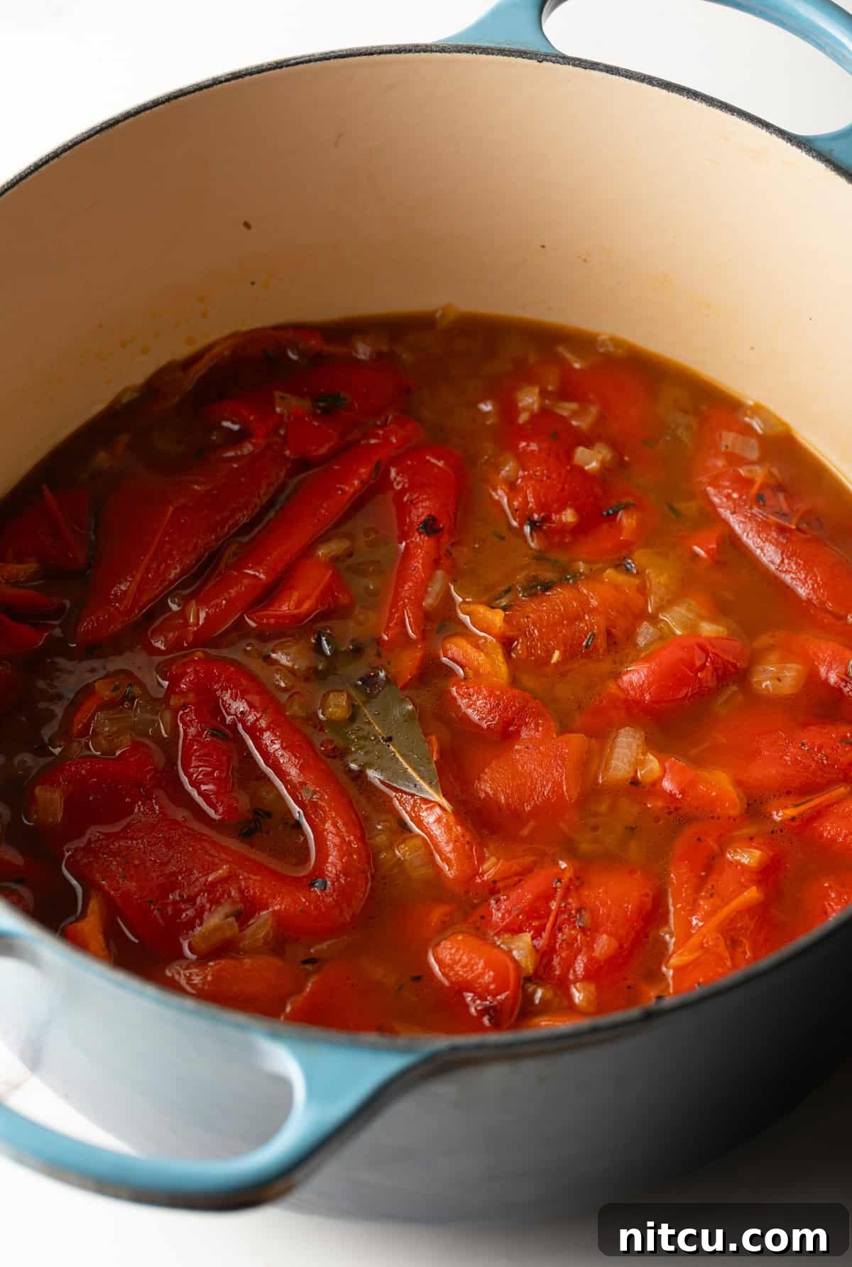 Peeled, tender roasted red peppers submerged in a rich, simmering broth with herbs, ready to be blended into a creamy soup.