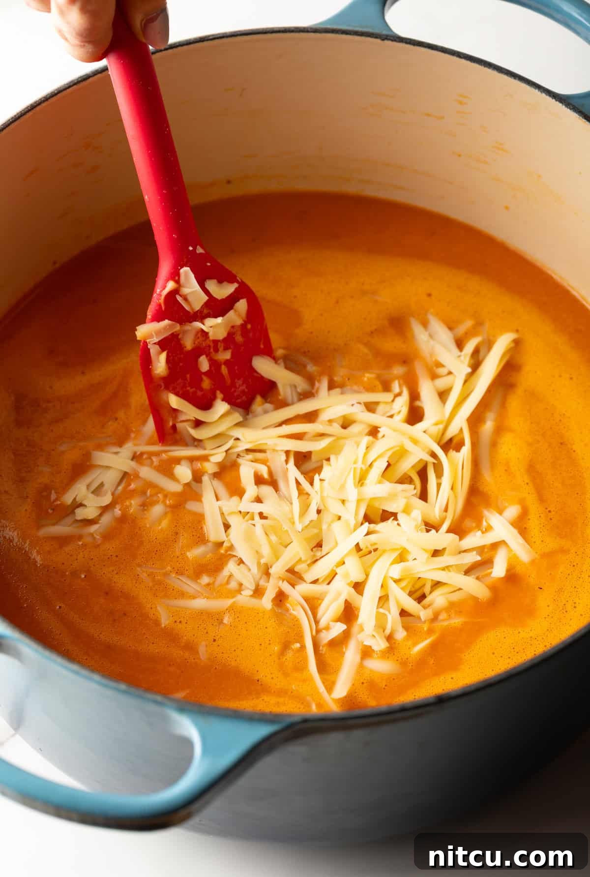 A generous handful of freshly shredded smoked Gouda cheese being stirred into a steaming pot of rich, creamy red pepper soup, ready to melt into a velvety texture.