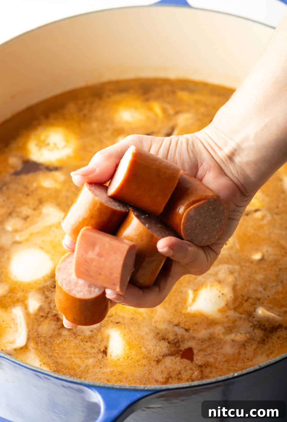 A hand carefully placing thick slices of kielbasa sausage into a steaming pot, joining the onions, potatoes, and fragrant Old Bay broth, simmering for the authentic Low Country Boil recipe.