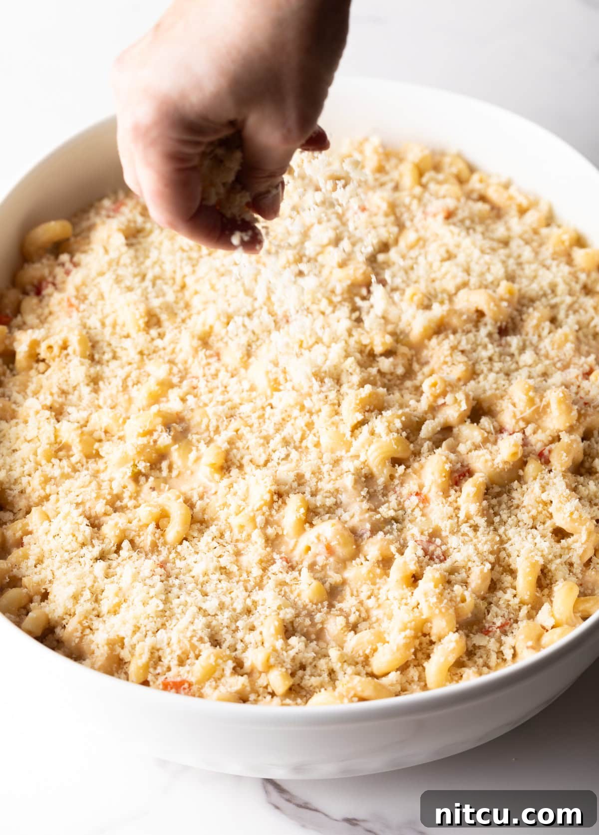 Breadcrumbs and grated parmesan are being sprinkled over the Cajun mac and cheese in a white baking dish.