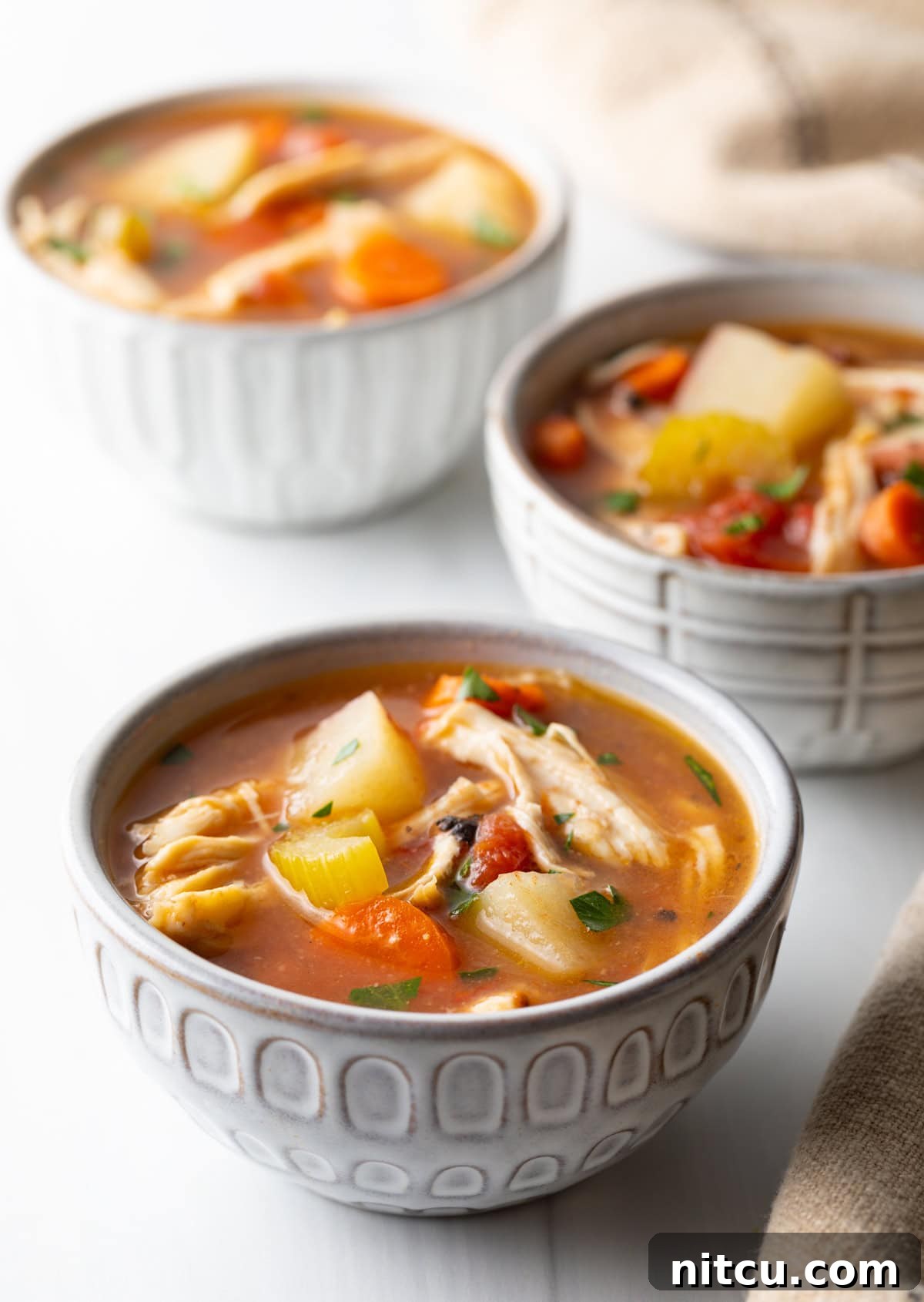 3 small white bowls of chicken stew with potatoes, carrots, and celery in a tomato broth.
