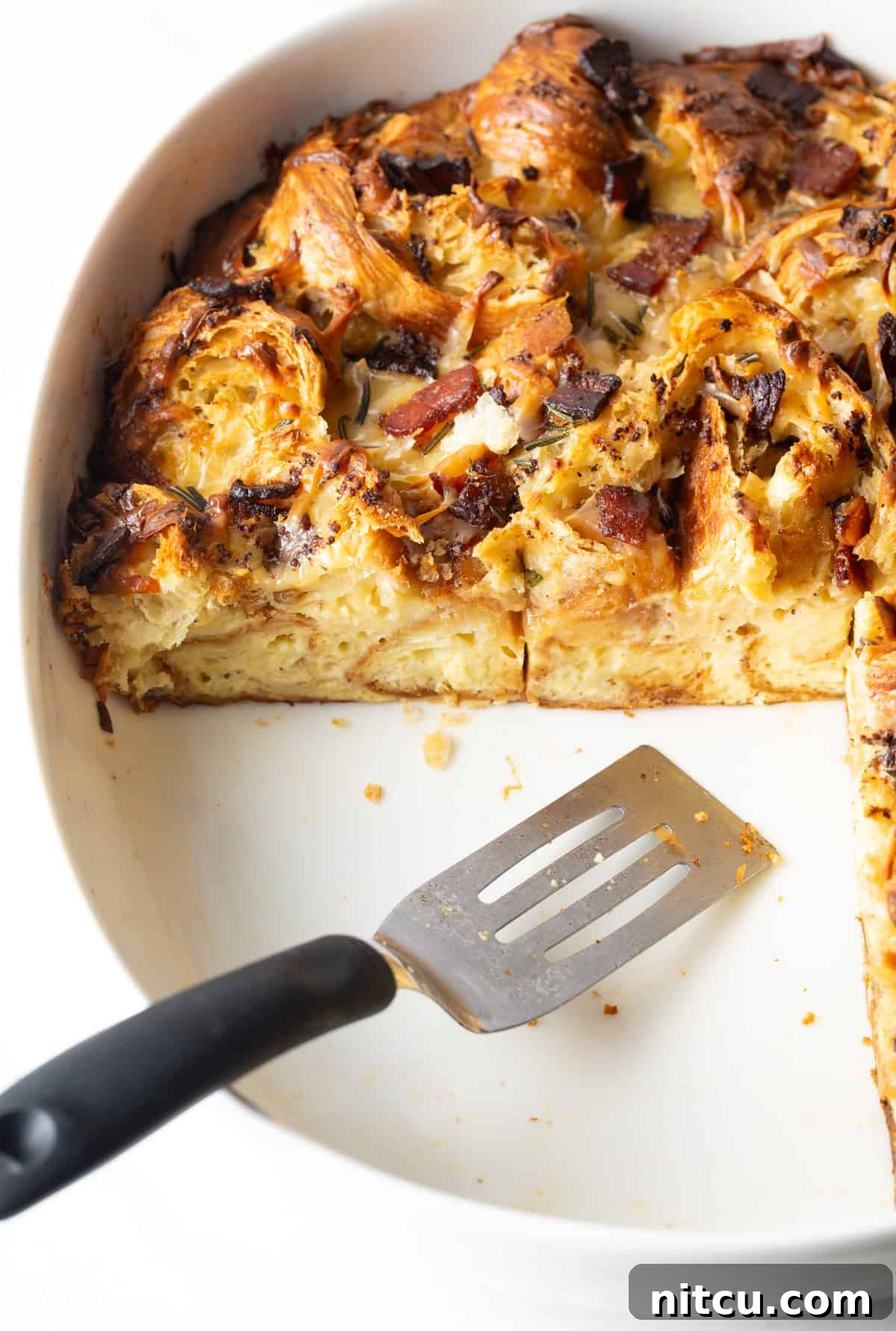 A metal spatula carefully serving golden slices of croissant casserole from a white baking dish onto plates.