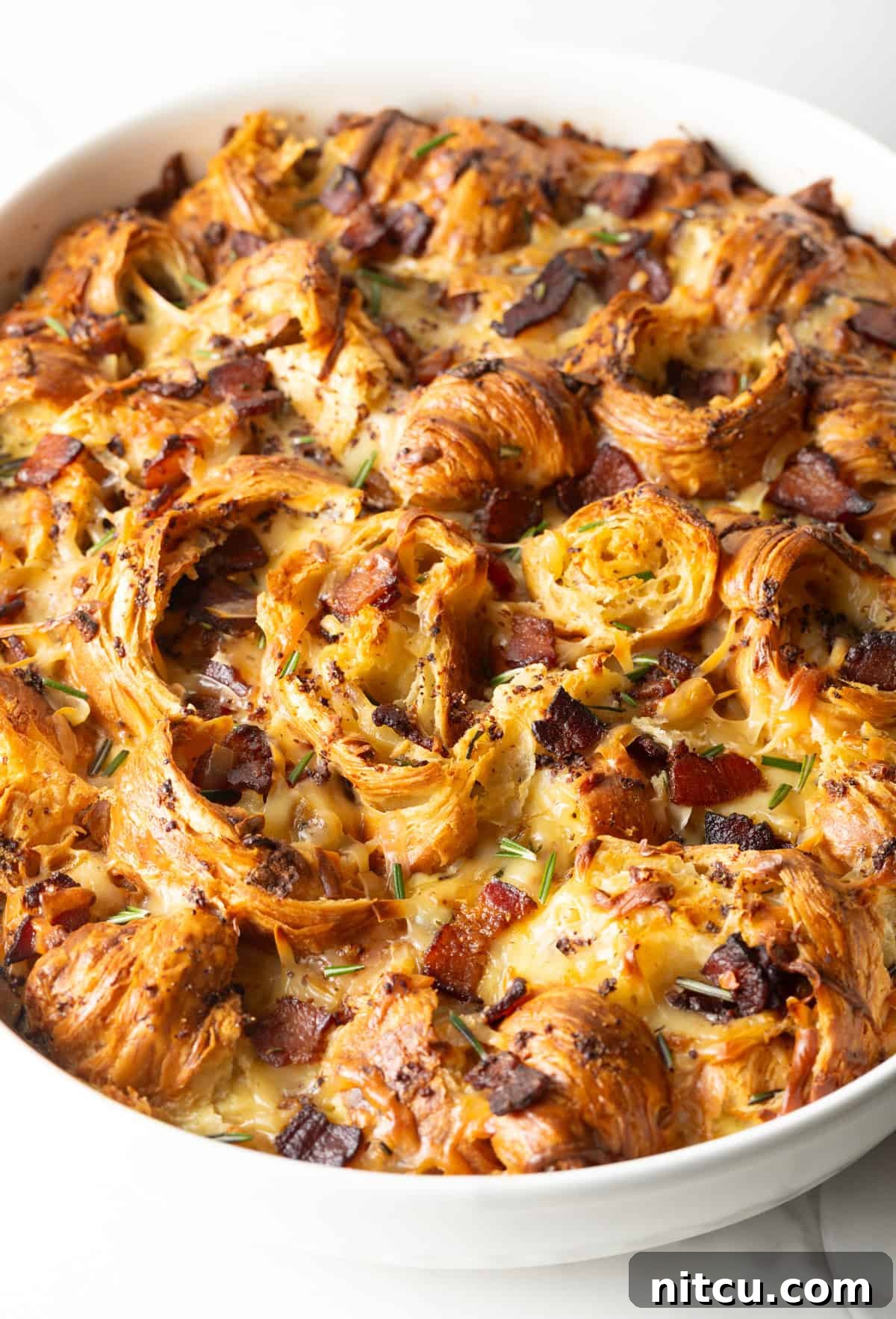 Close-up overhead shot of the croissant breakfast casserole in a white baking dish, showcasing its golden crust and delicious ingredients.