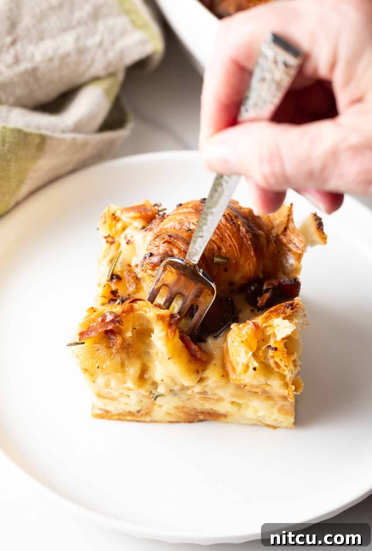 A fork taking a delicious bite out of a slice of warm croissant casserole, served on a white plate.