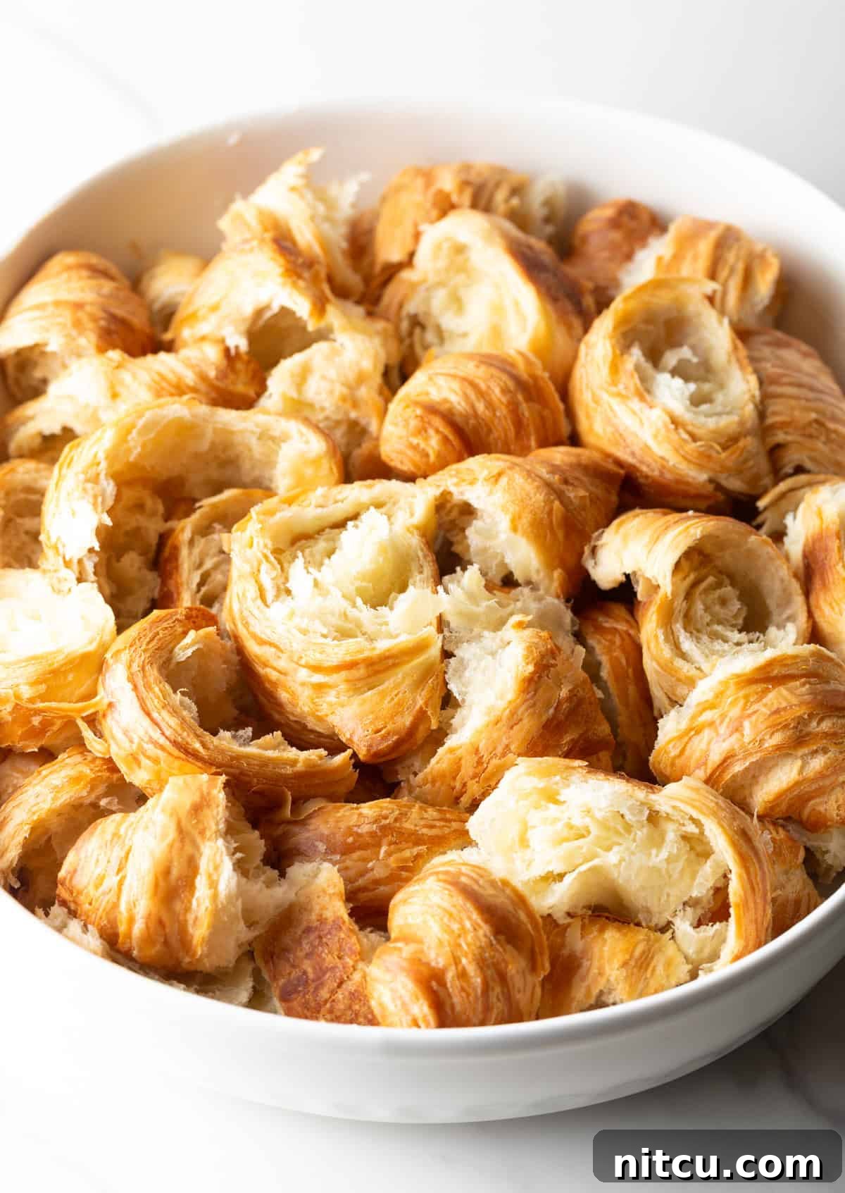 Torn croissants are meticulously layered in a white casserole dish, ready for the next ingredients.