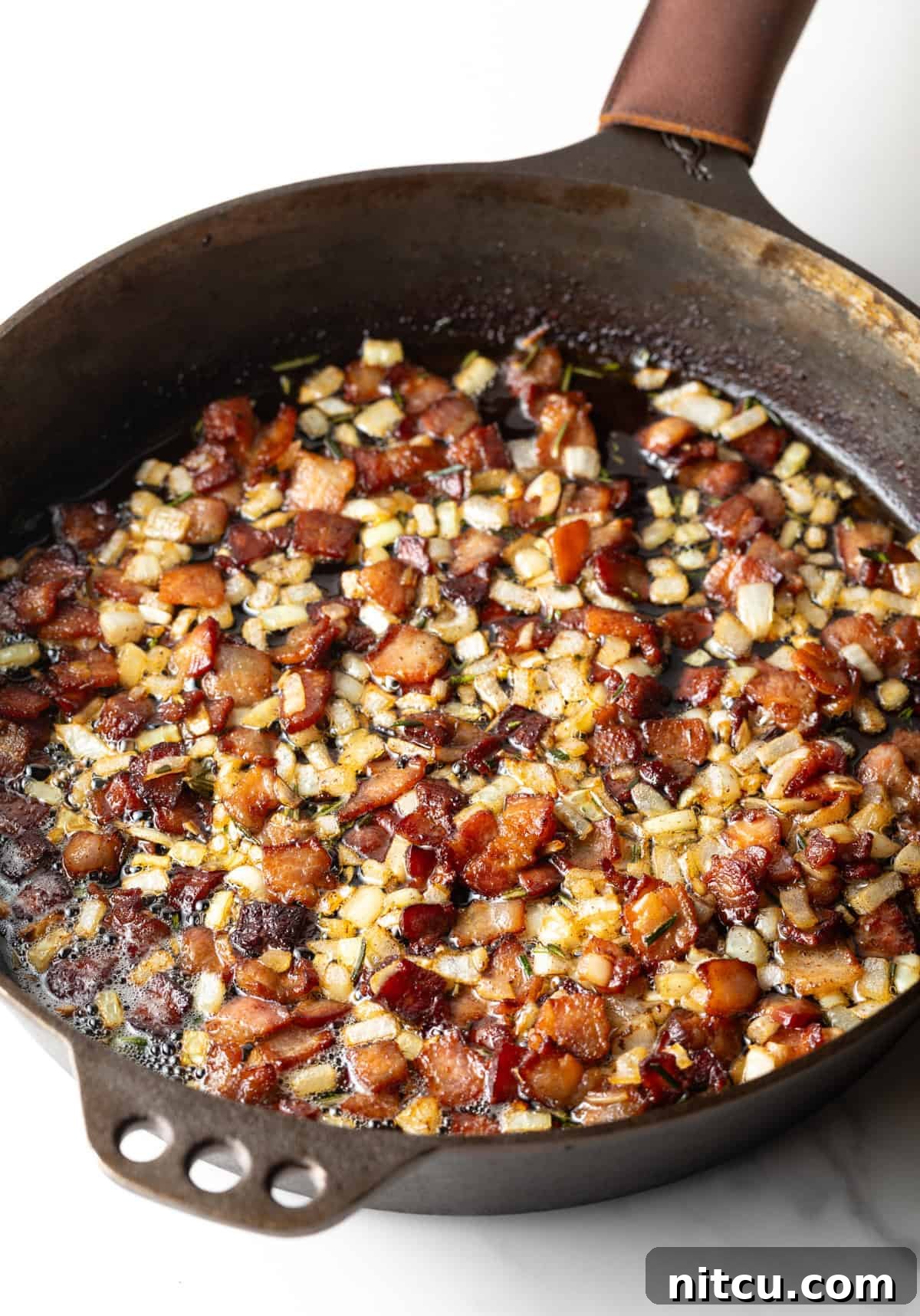 A black skillet filled with sizzling bacon, diced onions, and fresh rosemary being sautéed over medium heat.