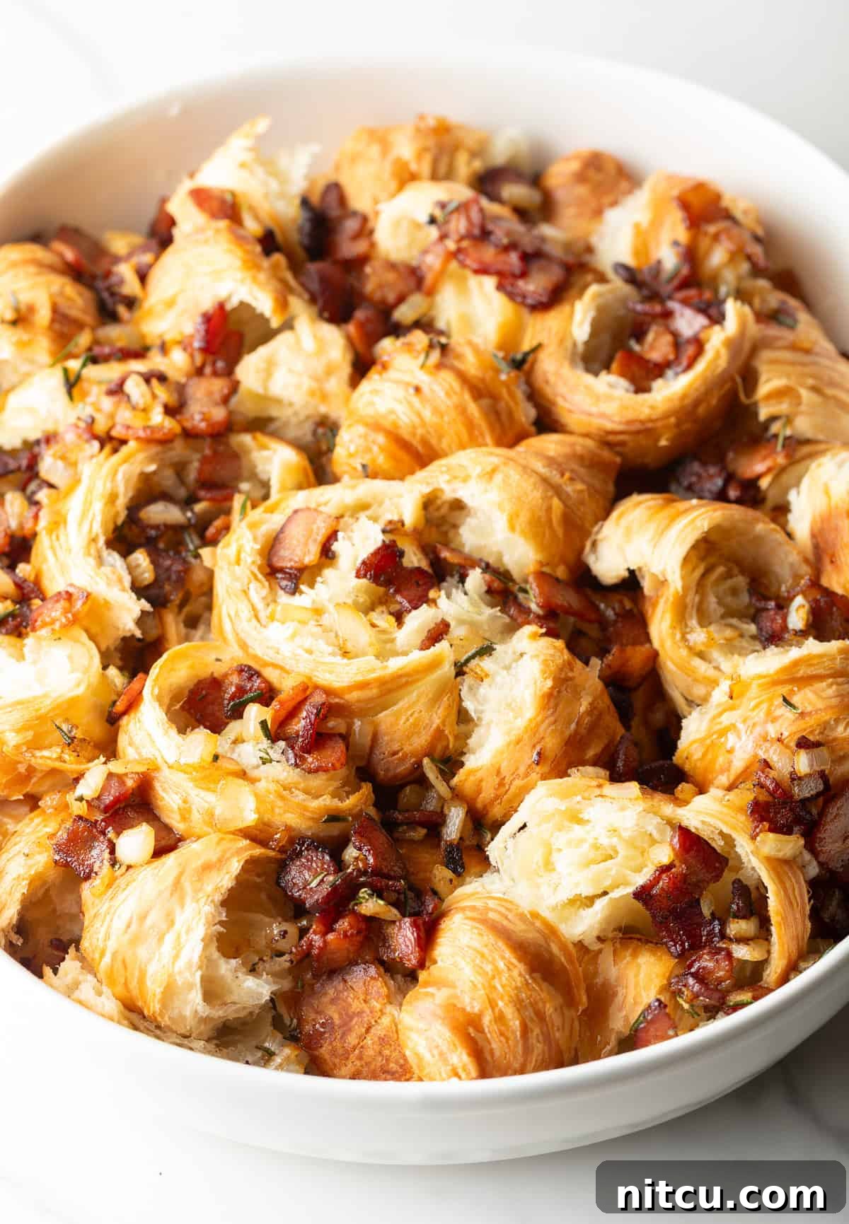 A savory bacon and onion mixture being carefully added with a slotted spoon to the base of torn croissants in a white baking dish.