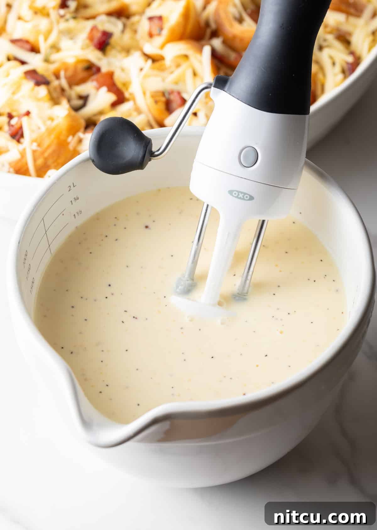 Creamy half & half, vibrant eggs, and coarse Dijon mustard being vigorously whisked together in a white mixing bowl.