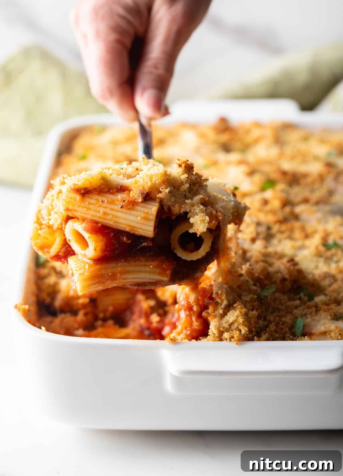 A serving of easy chicken parmesan casserole is being scooped out of a white baking dish, showing the layers of pasta, chicken, sauce, and cheese with a golden-brown crispy topping.