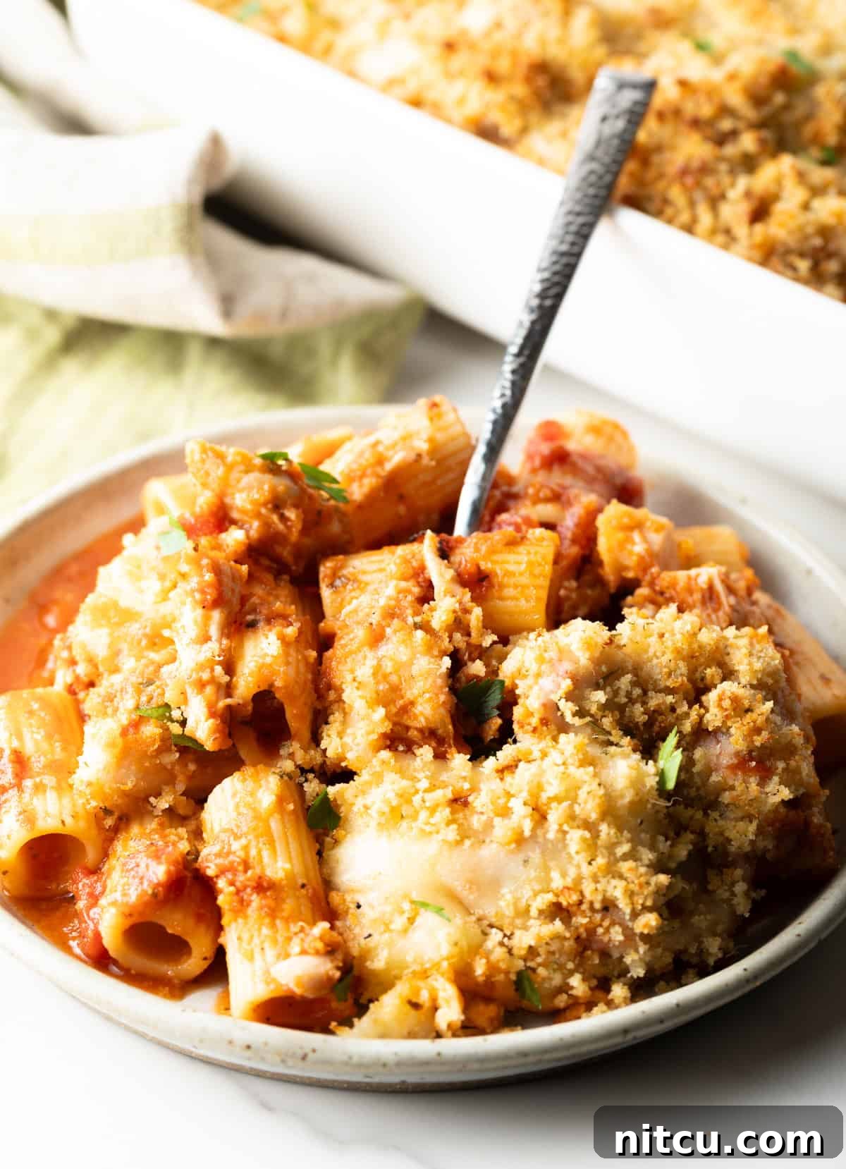 A fork lifting a bite of chicken parmesan casserole from a white plate, showcasing the melty cheese, chicken, and pasta.