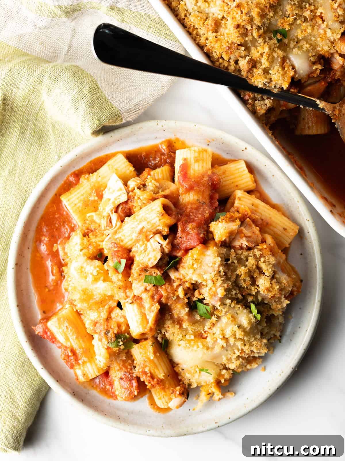 A serving of chicken parmesan casserole on a white plate, with a large white baking dish containing the rest of the casserole nearby.