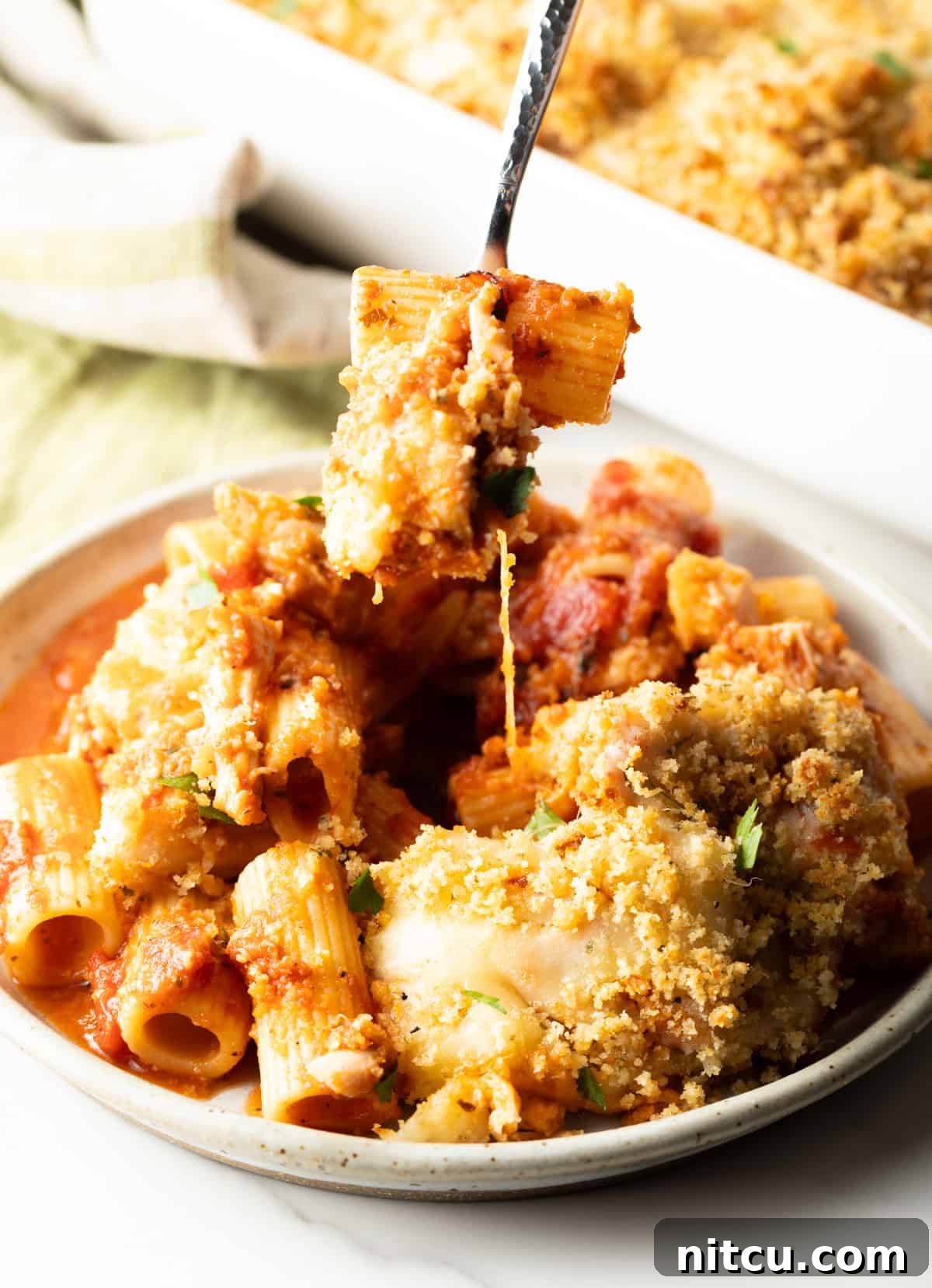 A fork taking a bite out of a serving of chicken parmesan casserole from a white plate, showing the melted cheese and pasta.