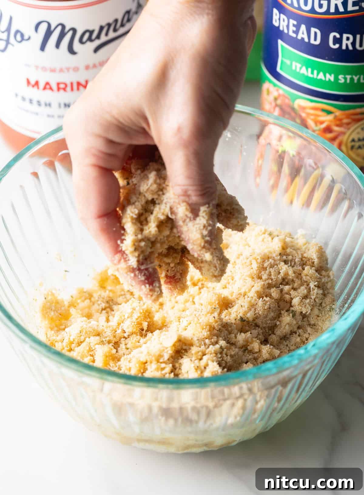A hand mixing a parmesan and breadcrumb mixture with melted butter in a glass bowl.