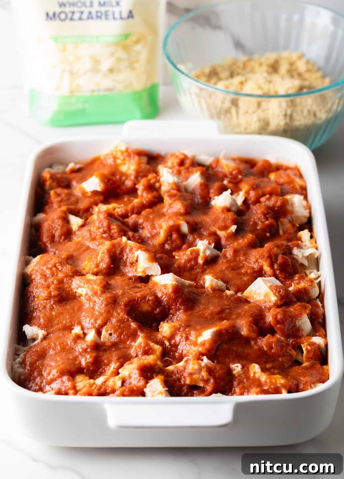 A white baking dish with cooked pasta, chopped chicken, and marinara sauce spread over it, ready for the next layers of chicken parmesan pasta bake.