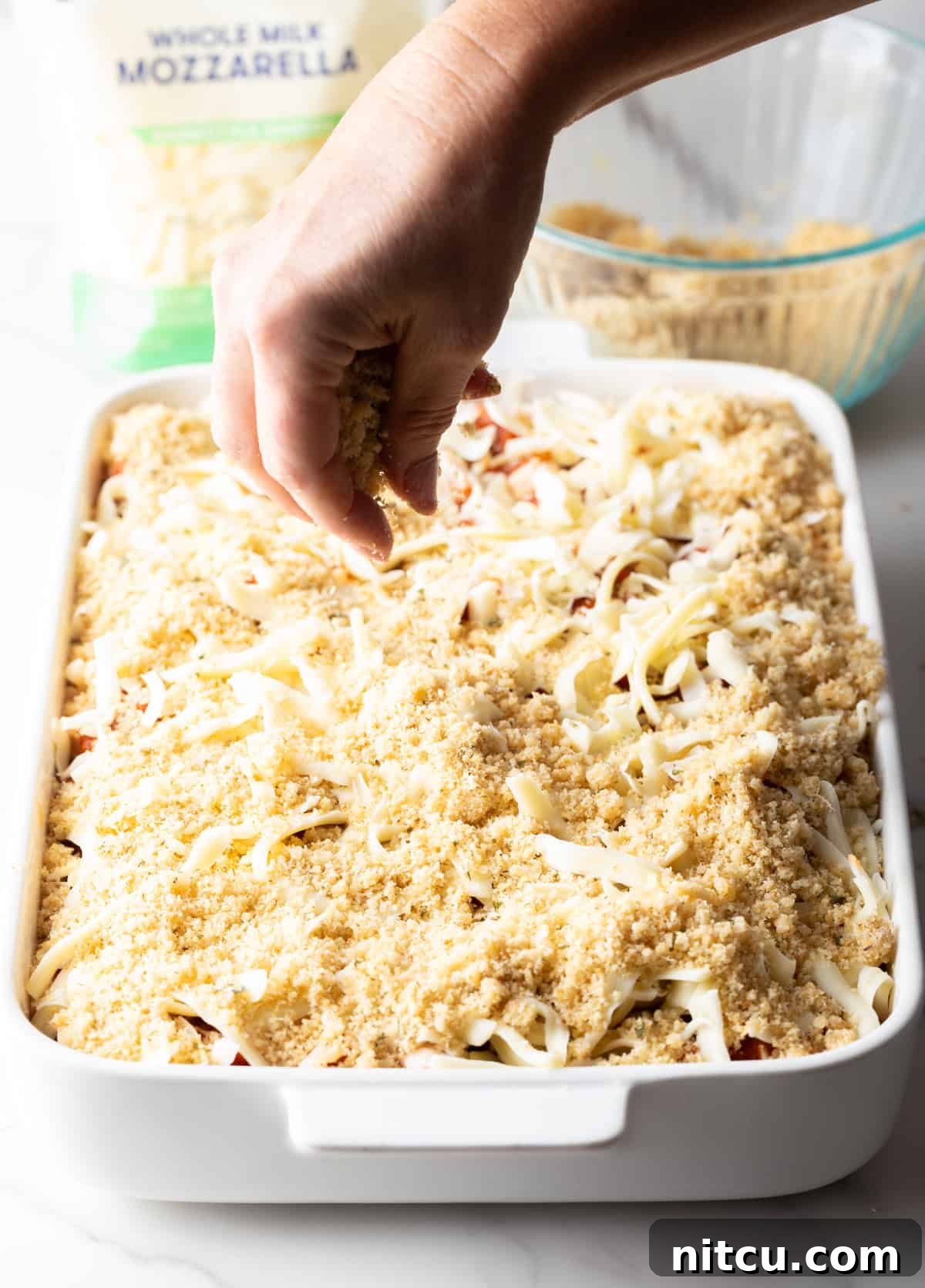 Shredded mozzarella cheese and the breadcrumb mixture being sprinkled over the chicken parmesan casserole layers in a white baking dish.