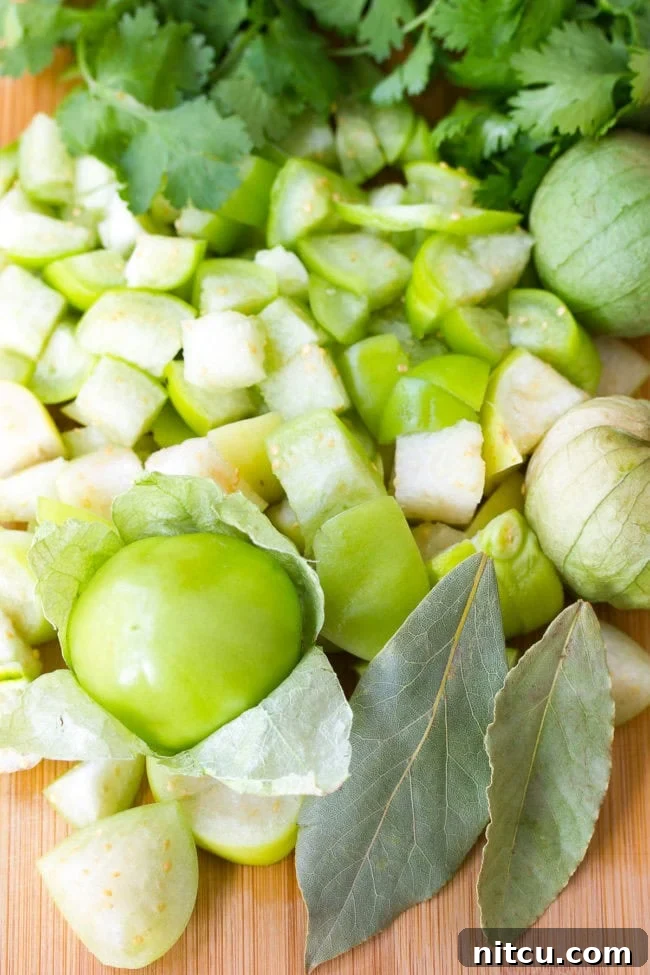 Diced tomatillos on a cutting board alongside fresh cilantro and bay leaves, prepared for a recipe.