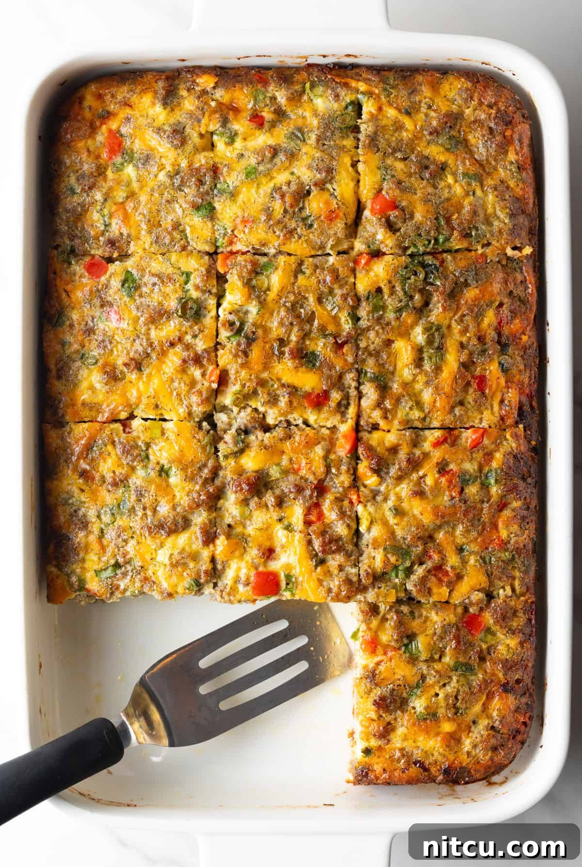 Pan of egg casserole with sausage and vegetables. A spatula is in the pan, with square portions of the casserole removed.
