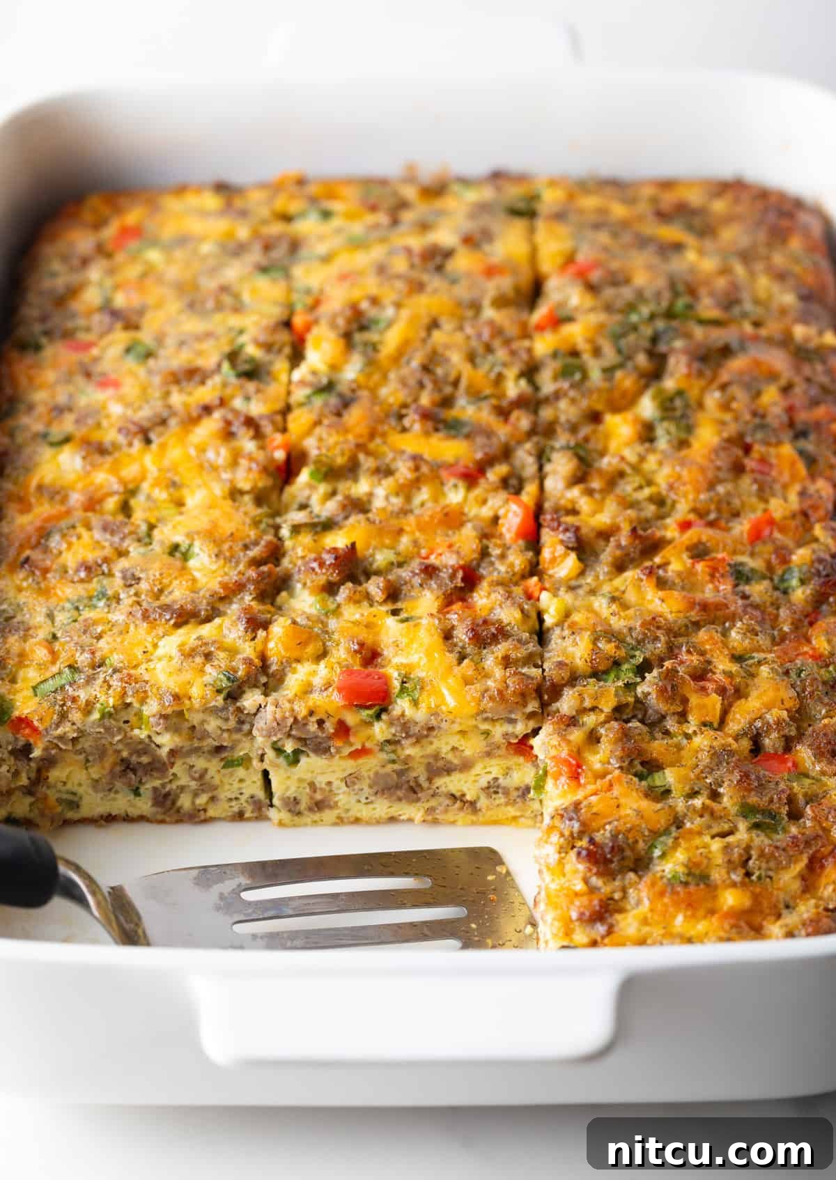 Pan of egg casserole with sausage and vegetables. A spatula is in the pan, with square portions of the casserole removed.