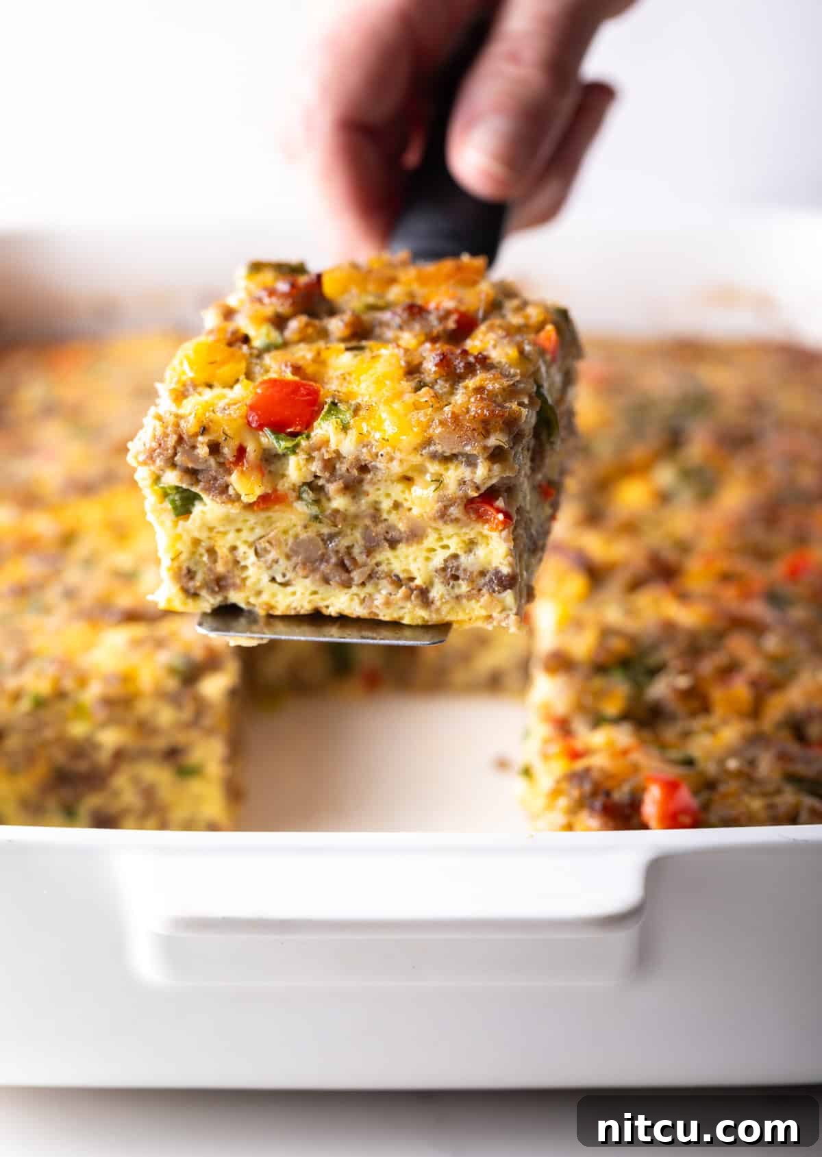 Pan of egg casserole with sausage and vegetables. A hand is holding a spatula above the pan, with a square portion.