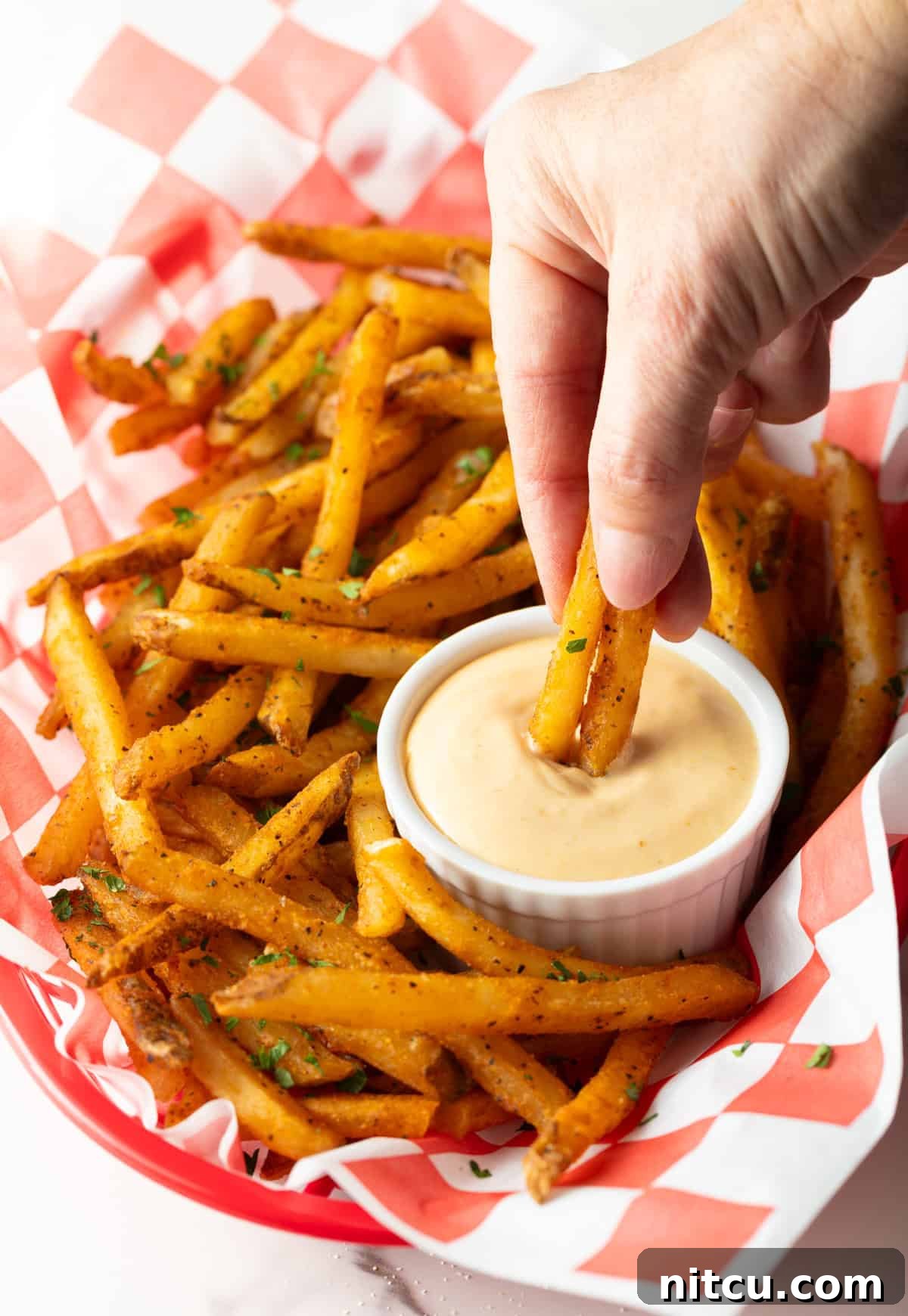 A basket of crispy French fries with a ramekin of French fry dipping sauce, and a hand is dipping two fries into the sauce.