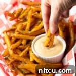 A basket of crispy French fries with a ramekin of French fry dipping sauce, and a hand is dipping two fries into the sauce.