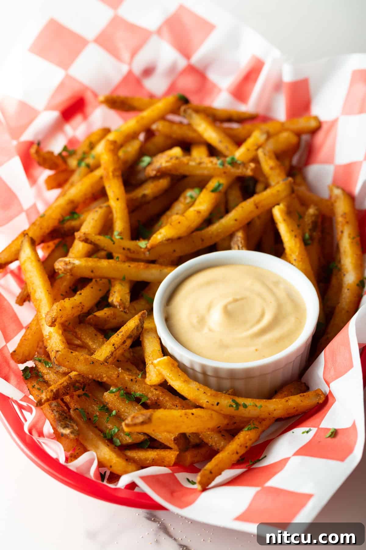A paper-lined basket of French fries surrounding a white ramekin filled with creamy French fry sauce.