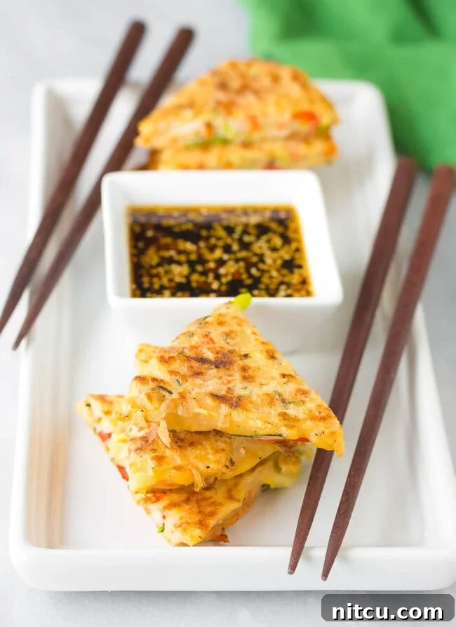Crispy Korean pancakes, Pajeon, cut into wedges and elegantly served on a plate next to a small bowl of spicy soy dipping sauce. The golden-brown edges promise a satisfying crunch.
