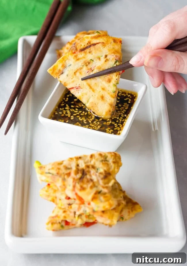 A hand is gently dipping a fresh, crispy Korean pancake wedge into a small bowl of spicy soy dipping sauce, highlighting the perfect pairing.
