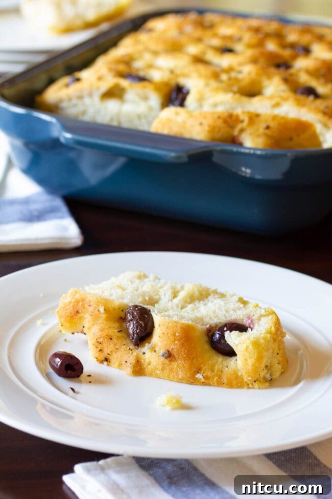  A loaf of focaccia is in a blue baking dish in the background. In the foreground is a white plate with a slice of fresh baked bread studded with olives.