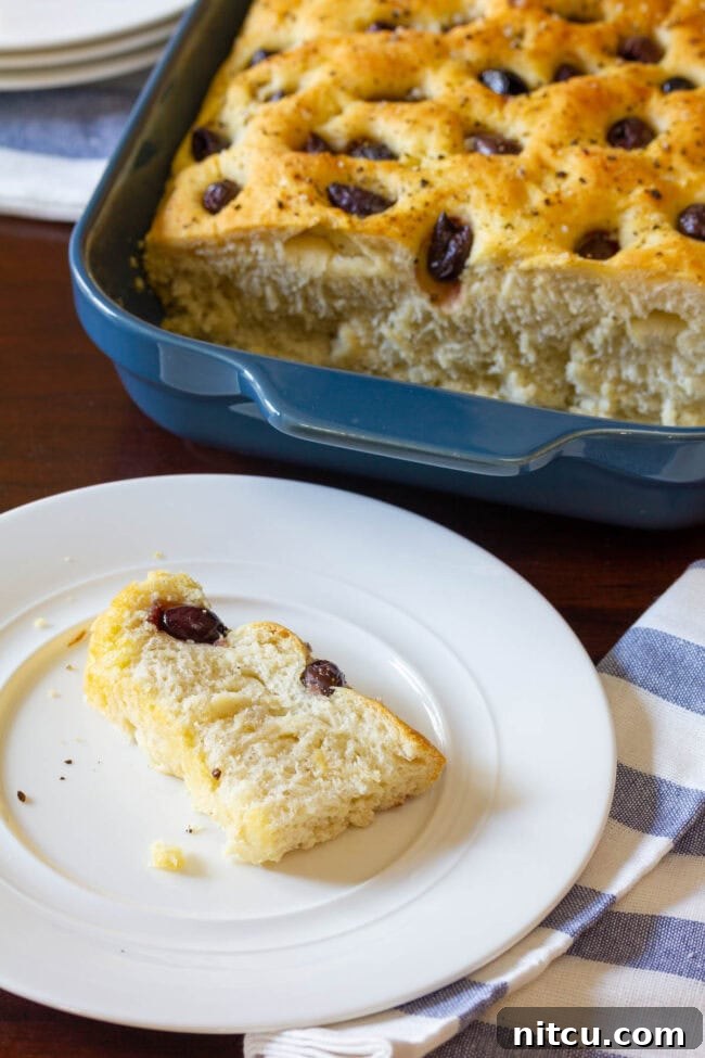  A loaf of focaccia is in a blue baking dish in the background. In the foreground is a white plate with a slice of fresh baked bread studded with olives.