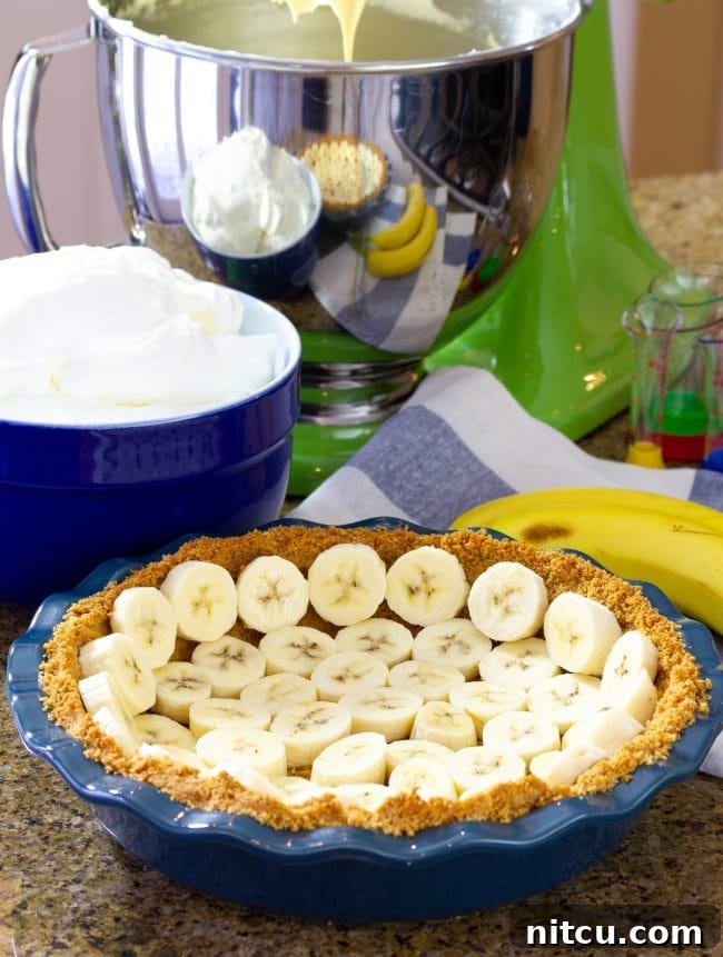 Freshly sliced banana pieces being arranged onto a baked graham cracker crust in a blue pie plate.