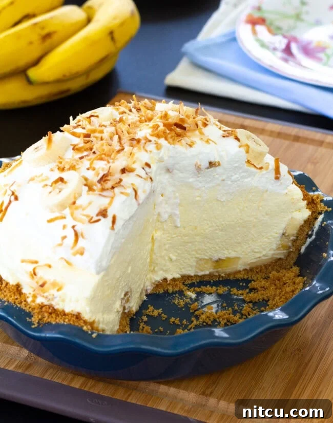 The finished banana cream pie in a pie pan, showcasing the golden graham cracker crust, a towering whipped cream topping, and toasted coconut sprinkles.