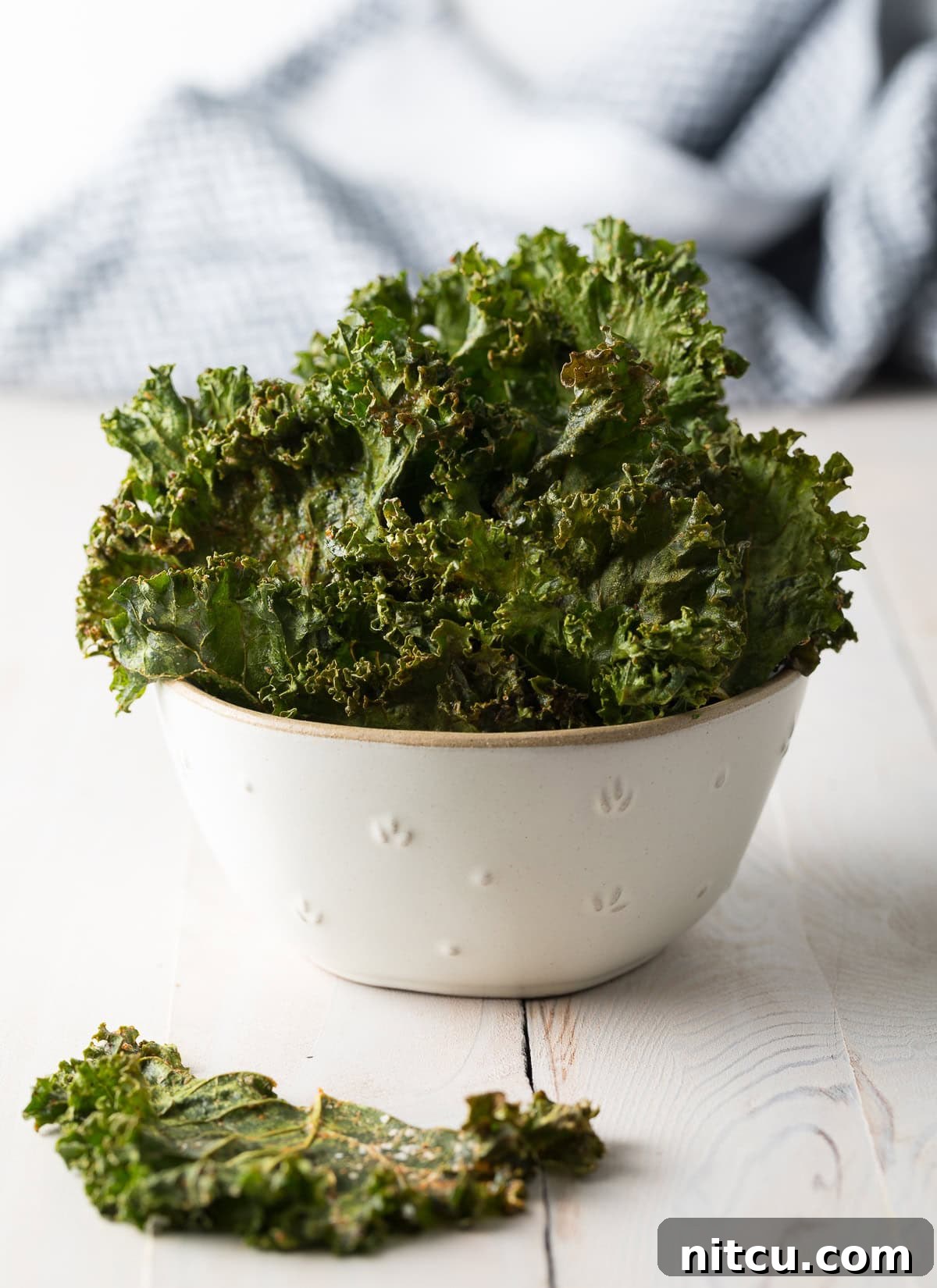 White serving bowl filled with crispy kale chips on a wooden surface.