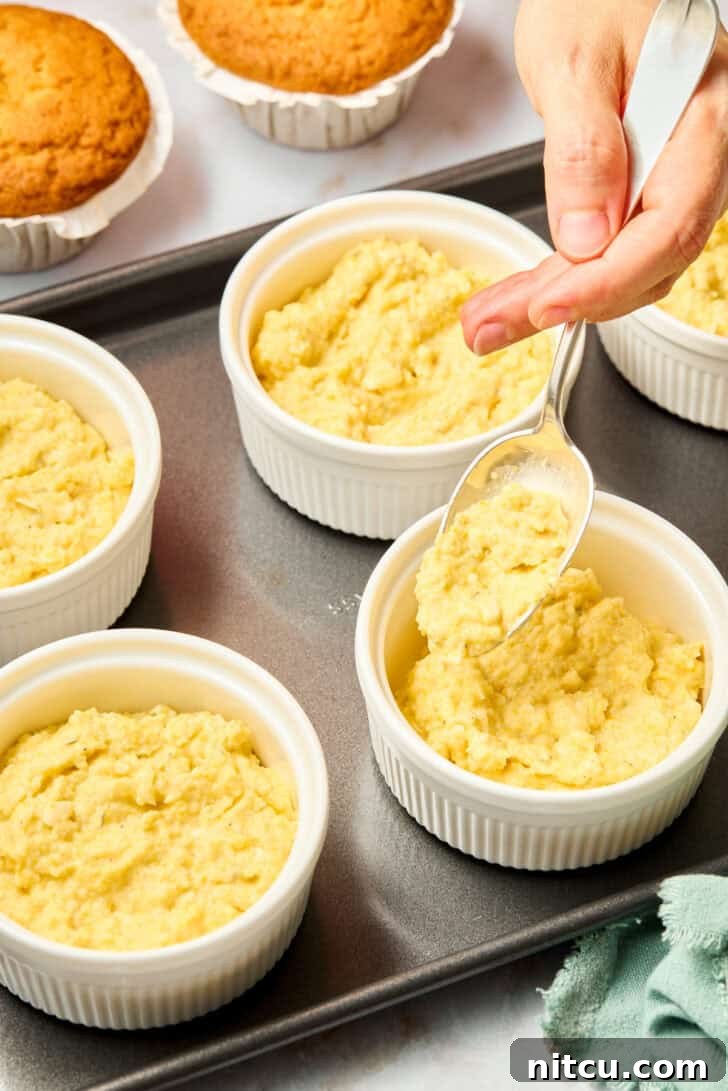 Hands pouring creamy Squash Casserole batter into individual ramekin dishes on a baking sheet