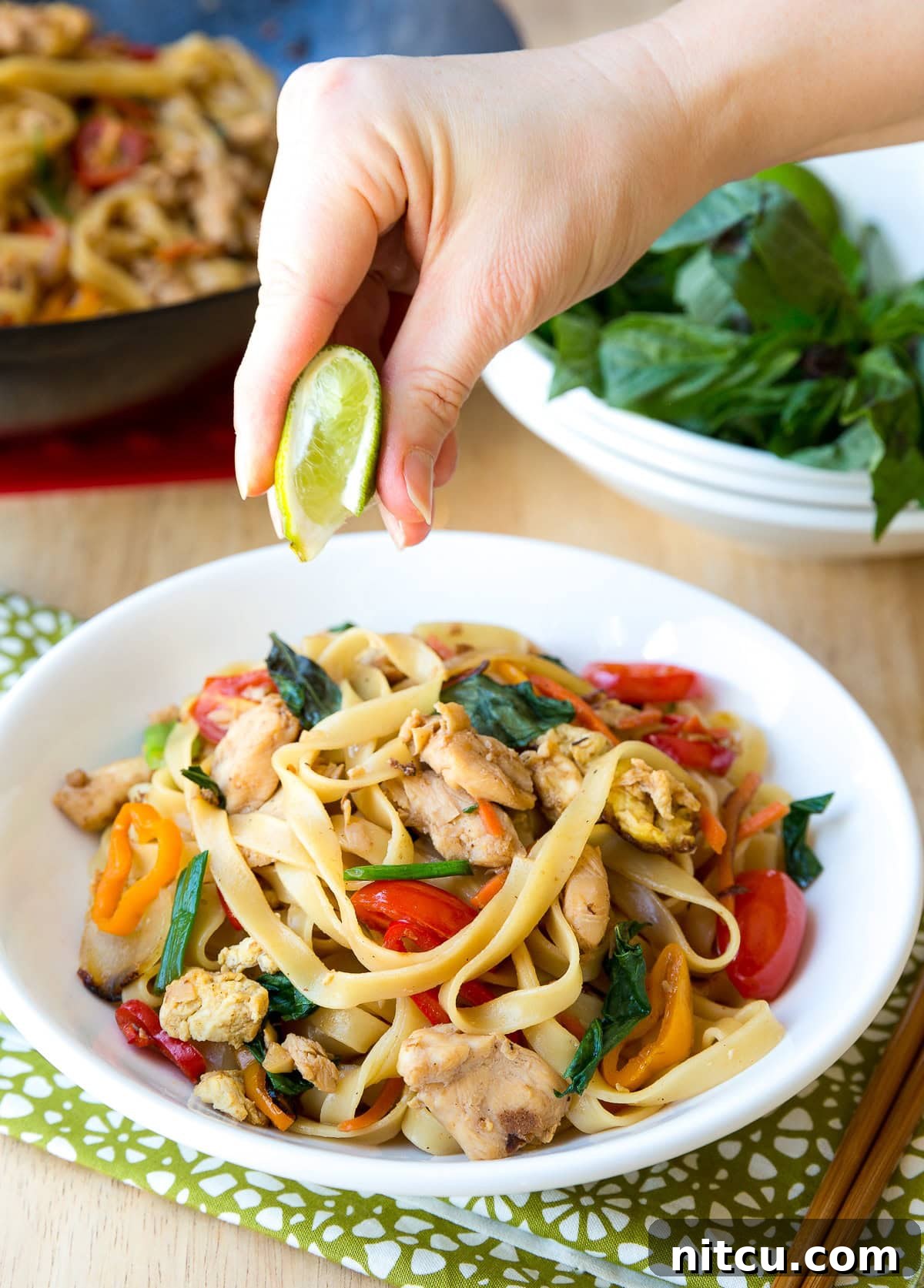 A hand squeezing a fresh lime wedge over a steaming bowl of Pad Kee Mao (Drunken Noodles), enhancing its fresh flavors.