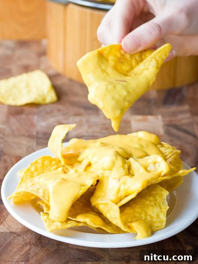 A hand taking a tortilla chip dipped in queso from a plate of queso-topped chips, showcasing the creamy texture.