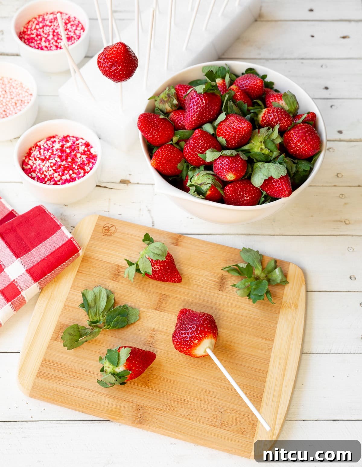 A top-down view showing a white bowl filled with fresh, whole strawberries. Beside it, a wooden cutting board displays a few strawberries, one of which has its green top removed and a lollipop stick carefully inserted through its center, ready for dipping.