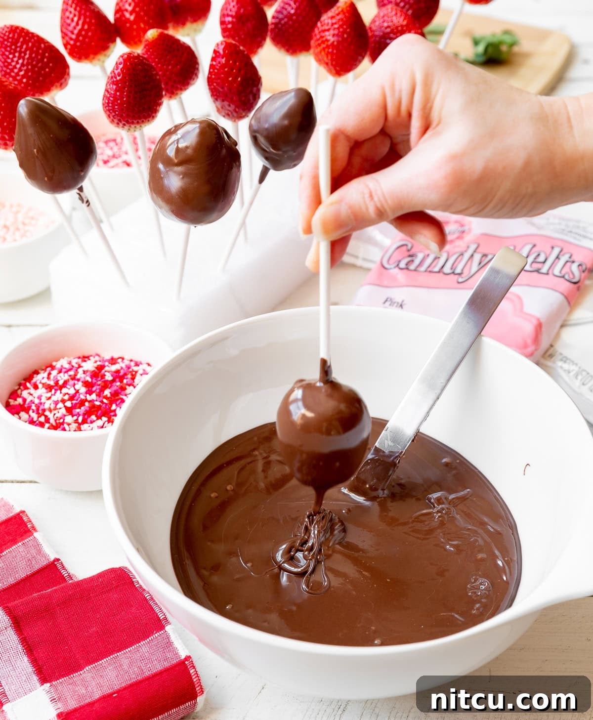 A close-up shot of a hand gently dipping a fresh strawberry, impaled on a lollipop stick, into a bowl of perfectly melted brown chocolate. The chocolate smoothly coats the berry, signaling the beginning of its transformation.