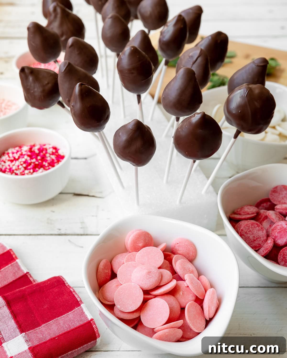 A styrofoam block serves as a drying rack, holding a dozen meticulously crafted chocolate-covered strawberry lollipops. Around the base of the block are several white bowls, neatly arranged with vibrant pink, rich red, and creamy white melted candy melts, alongside a bowl of cheerful pink and white sprinkles, ready for decoration.