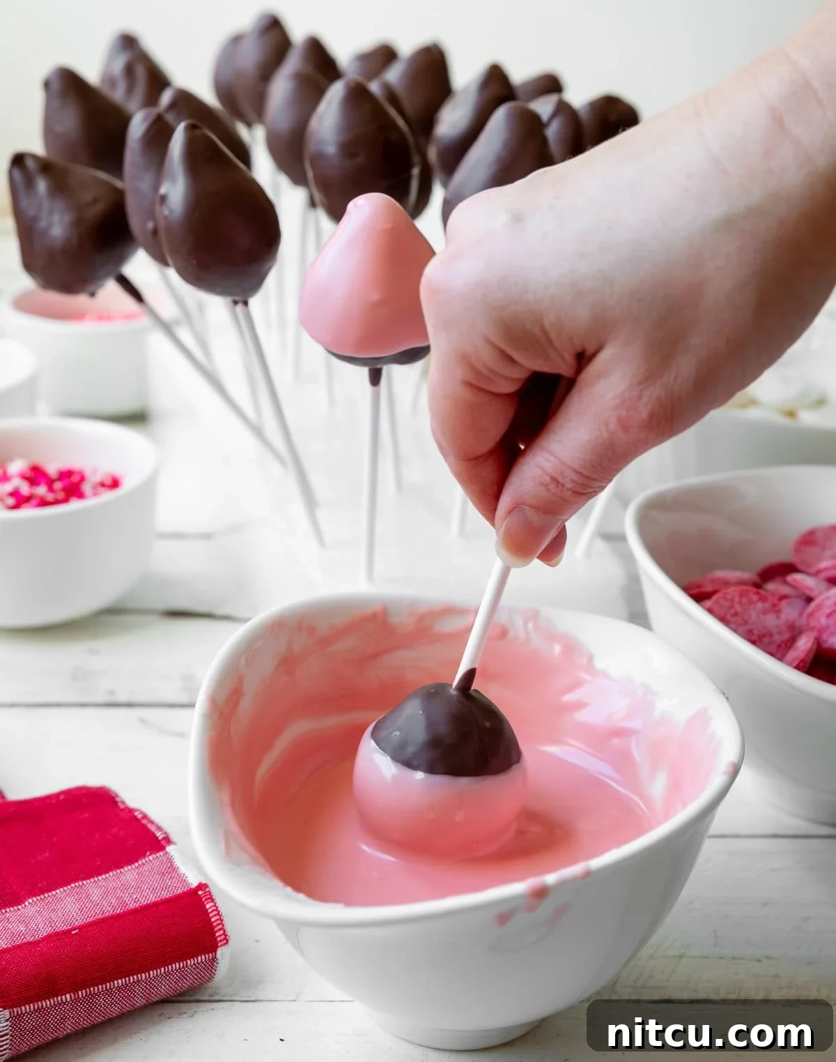 A hand carefully dips a brown chocolate-covered strawberry pop into a bowl of melted pink chocolate, creating a beautiful layered effect. This step adds vibrant color and another dimension of flavor to the treat.