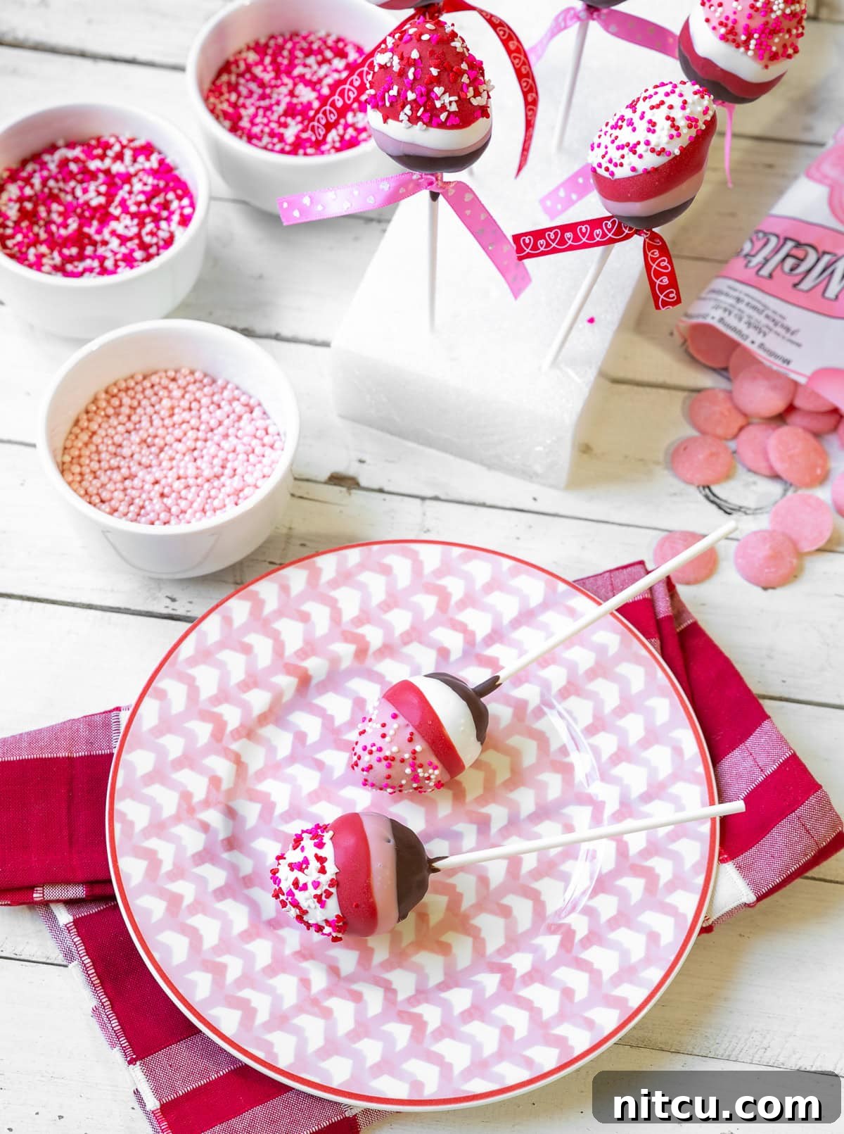 Two exquisite chocolate-covered strawberry lollipops, beautifully layered with rich brown chocolate, delicate pink, pure white, and vibrant red, all artfully topped with charming pink and white Valentine's Day sprinkles. They are presented against a soft, romantic background.