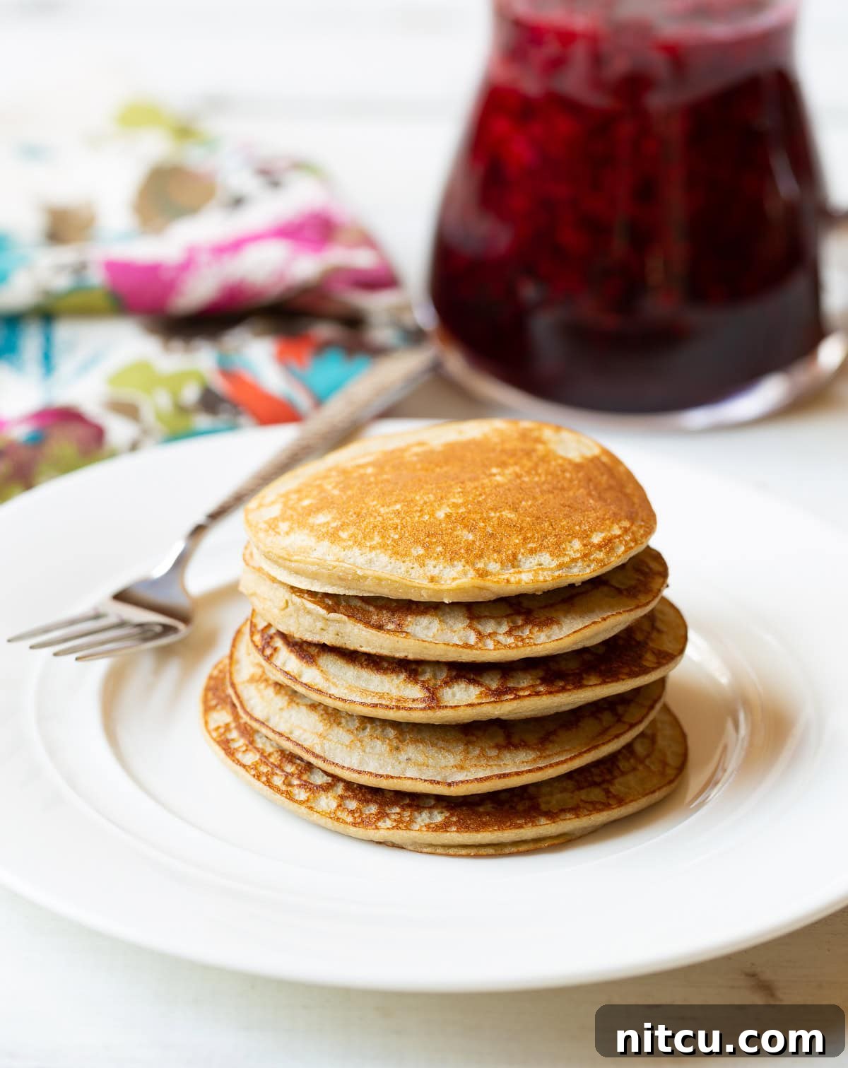 A beautifully stacked tower of five fluffy paleo pancakes, perfectly golden brown, resting on a pristine white plate, invitingly ready to be enjoyed.