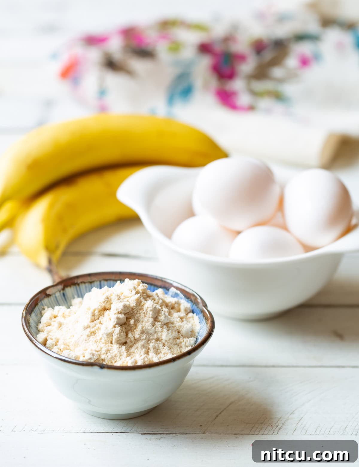 Essential ingredients for paleo pancakes: a small bowl of white coconut flour, six fresh eggs in a bowl, and a bunch of ripe bananas, ready for blending.
