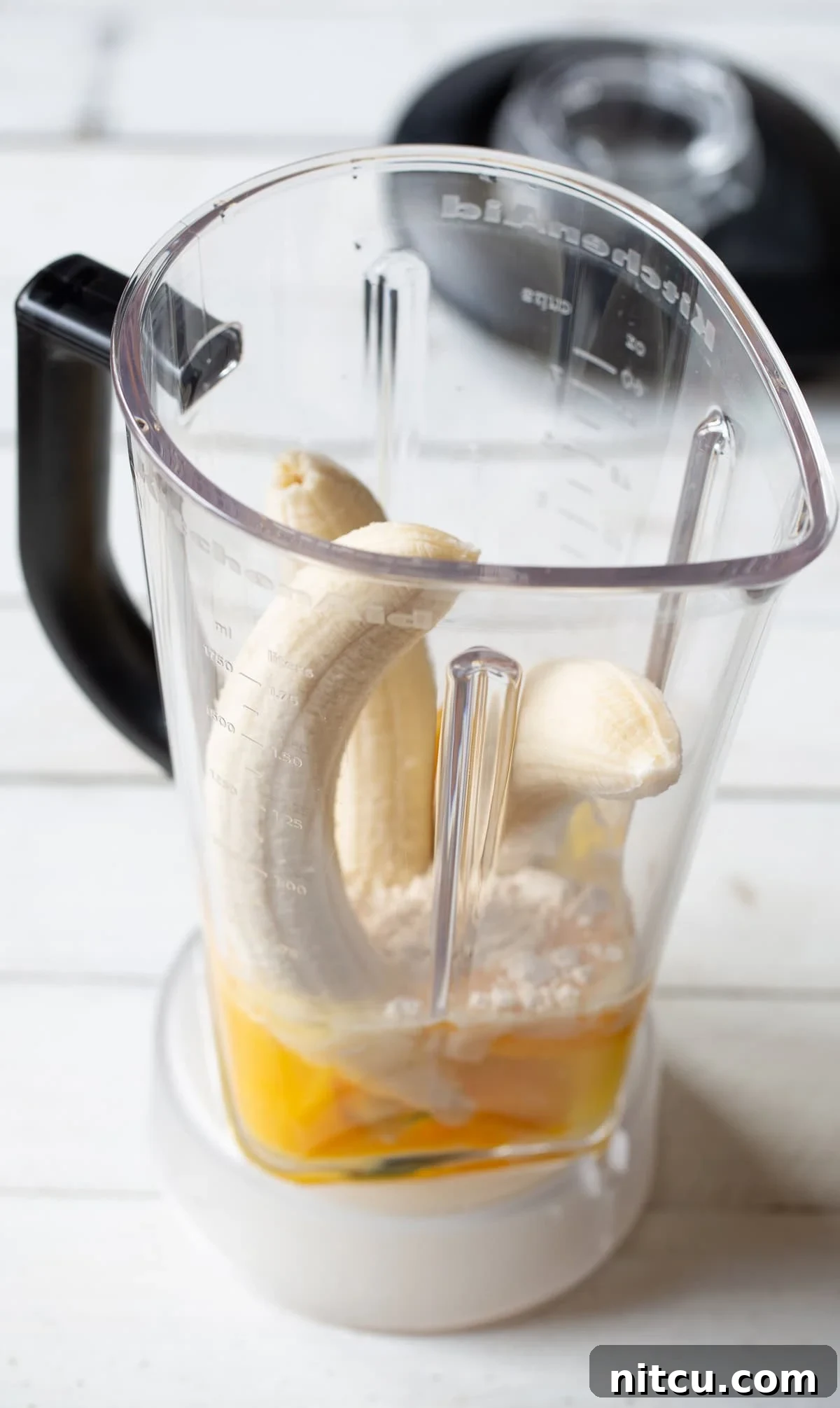 Three ripe bananas, a bowl of coconut flour, and eggs arranged inside a blender, ready to be combined into a smooth paleo pancake batter.