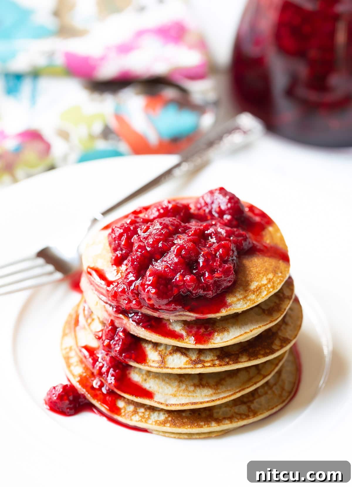 A stack of five perfectly cooked coconut flour pancakes, generously topped with vibrant red raspberry syrup, presented on a clean white plate.